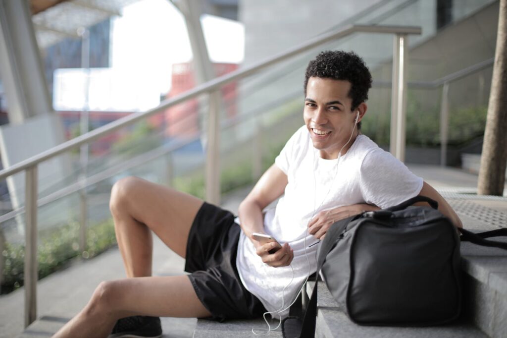 pexels-photo-3799268 Delighted African American man in earphones and sportswear smiling at camera while sitting with big black bag on stairs and listening to music against blurred exterior of contemporary building in city